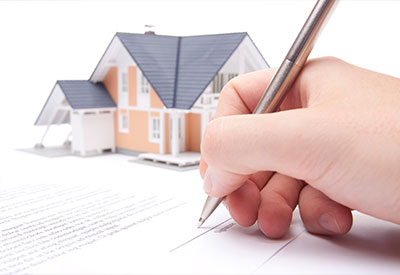 A hand holding a pen is poised to sign a property deed. In the background, a small model of a house with a blue roof hints at estate planning or activities related to real estate and home buying.