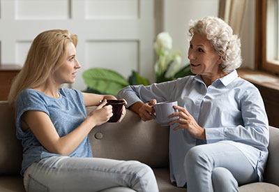 Two women sitting on a couch, smiling and holding mugs, seem to be engaged in a pleasant conversation about estate planning.