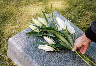 A hand gently places a bouquet of white lilies on a gray stone slab, surrounded by grass, symbolizing a serene farewell as part of thoughtful end-of-life planning.