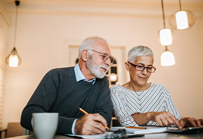 An older couple sits at a table, engaging in estate planning while looking at documents. The man holds a pencil, and the woman points to a paper.