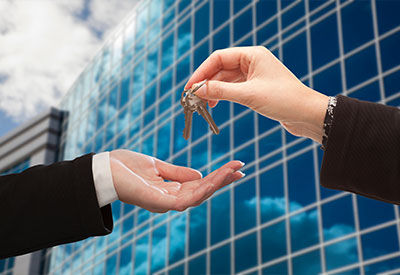 A pair of hands is shown against the backdrop of a glass office building and cloudy sky. One hand holds out a palm, while the other hand is giving a set of keys, symbolizing business succession and the careful transfer of legacy planning.