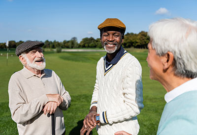 Three older men are standing on a golf course discussing business succession, smiling and chatting under the clear blue sky that casts a sunny glow over the green grass.