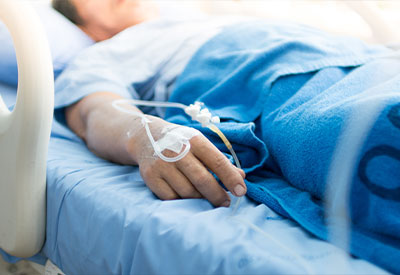 A person is lying in a hospital bed under a blue blanket, their hand visible with an IV drip attached