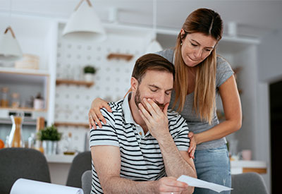 A man and woman are in a modern kitchen, looking concerned while reading a document about debts. The man, in a striped shirt, holds the paper and rests his head on his hand, while the woman, in a gray shirt, stands close with a supportive arm around his shoulder.
