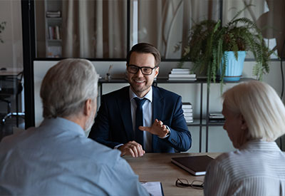 A young man in a suit and glasses is seated at a desk, smiling and gesturing as he discusses estate planning with an older couple sitting across from him. The office setting includes plants and bookshelves in the background.