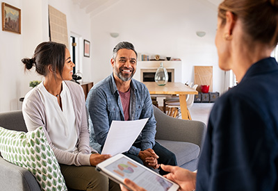 A couple meets with a financial advisor or estate planner in a home setting, discussing documents and viewing information on a tablet.