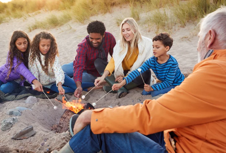 Family and friends roasting marshmallows over a campfire on a sandy beach.