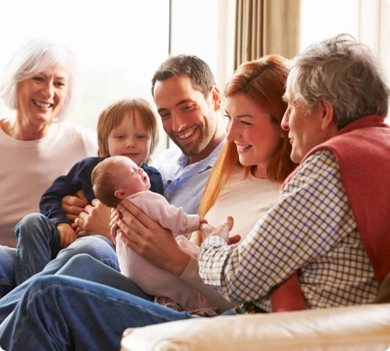 Happy family sitting on a couch, with grandparents, parents, and children smiling and interacting with a newborn baby.