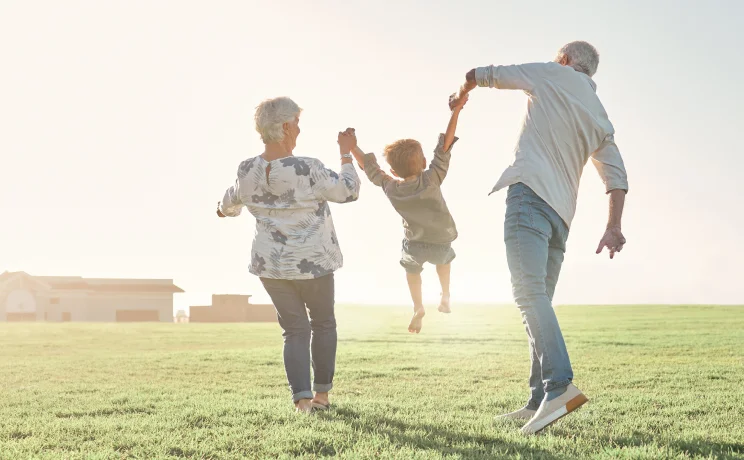 Grandparents joyfully swinging their grandchild by the arms while walking in a sunny, open field.